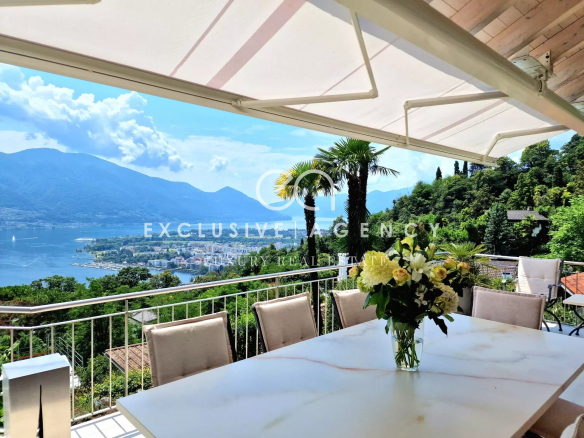 Outdoor terrace dining area with a white marble table and a bouquet of white flowers, overlooking a lake and mountains under a canopy.