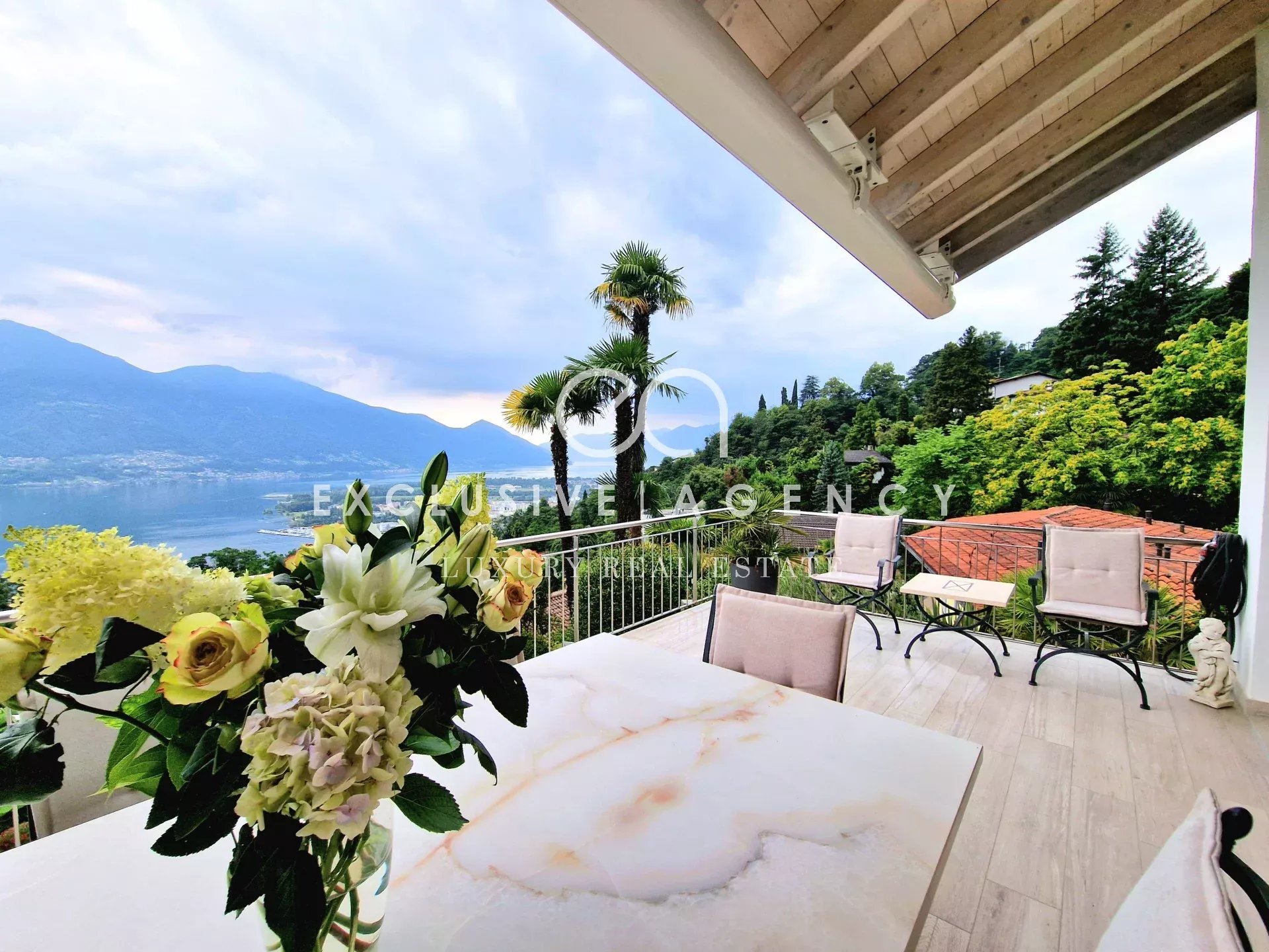 Balcony seating area with a marble table and a bouquet of white flowers, overlooking a lake and green hills. beige chairs surround the space.