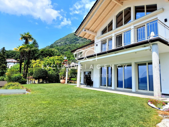 Modern white villa with a glass sliding-door facade, second-floor balcony, and a neatly mowed lawn with mountain backdrop.”] ,
