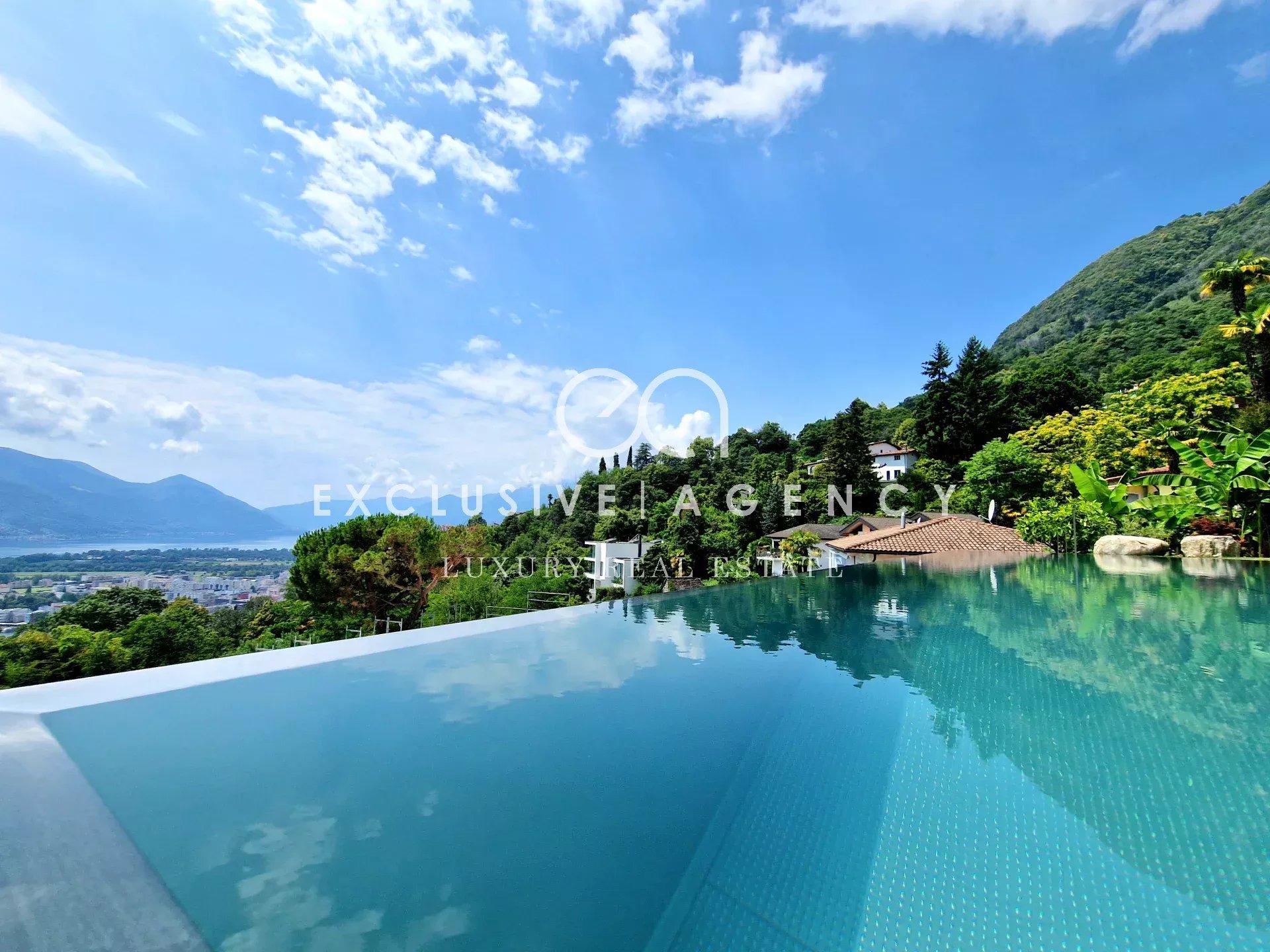 Infinity pool with turquoise water reflecting a blue sky, overlooking green hills and a hillside villa village.