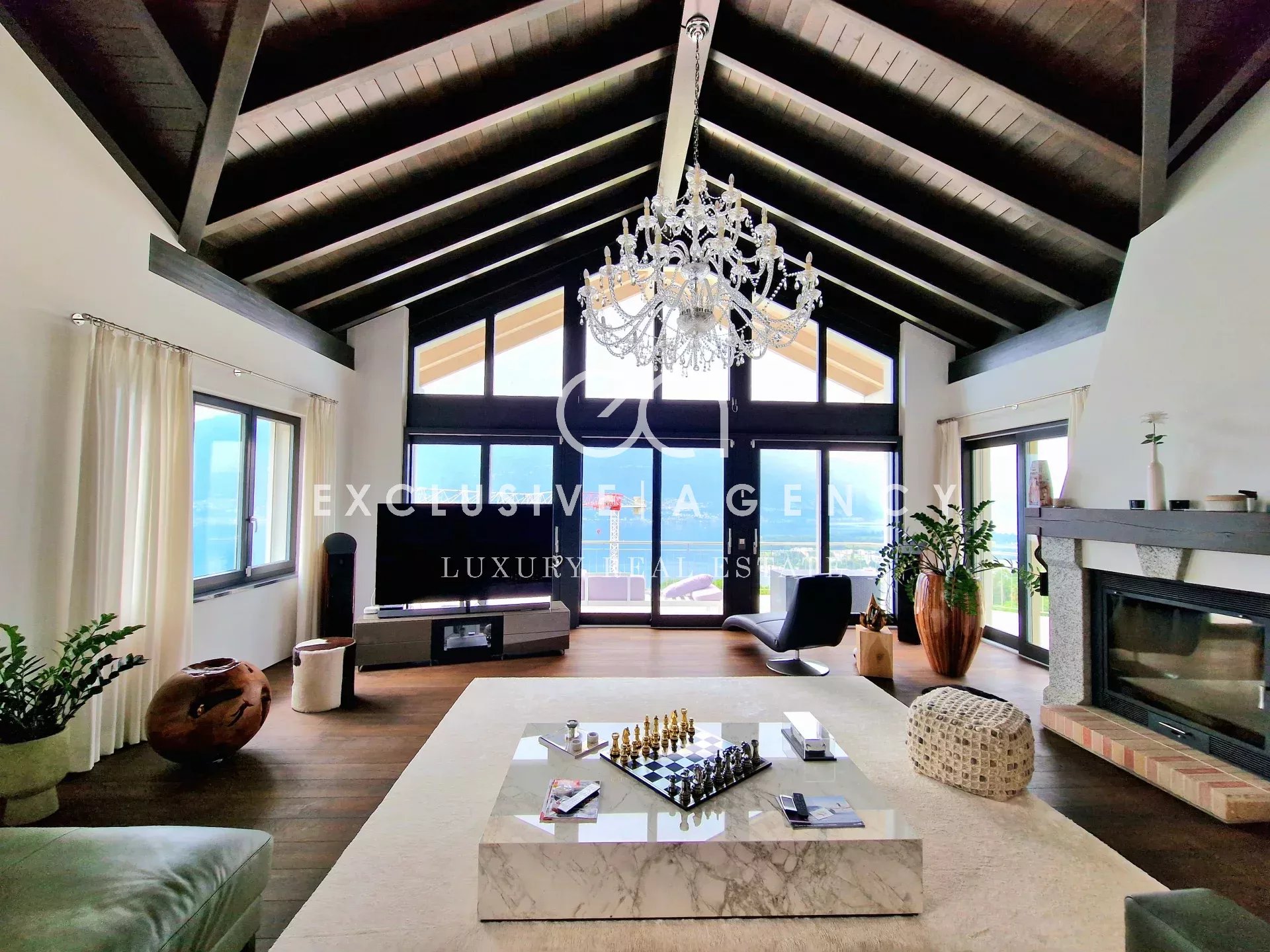 Luxurious living room with dark vaulted beams, crystal chandelier, and large glass doors opening to a view, centered by a marble chess-table coffee table.