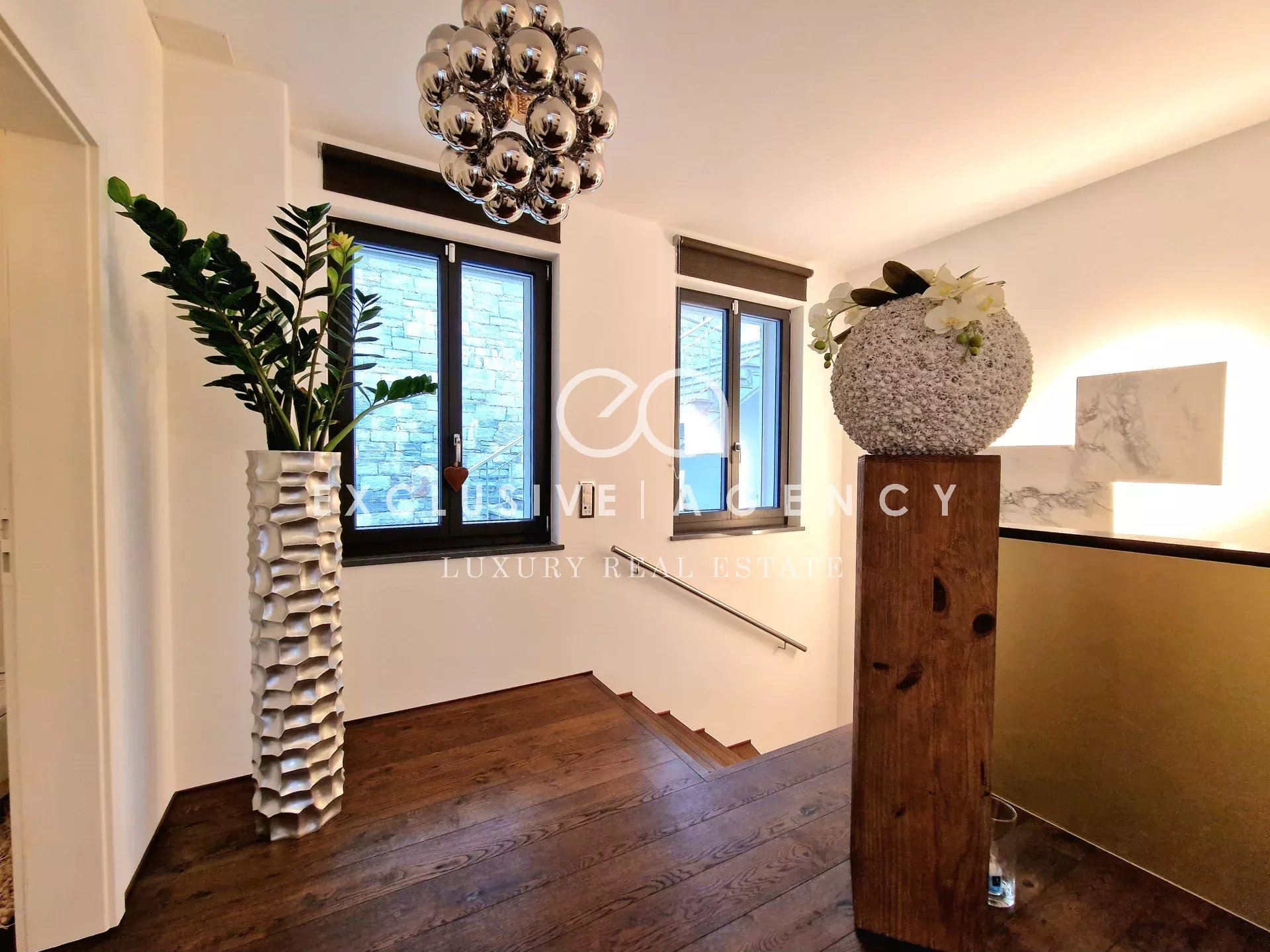 Luxurious stairwell with dark wood steps, a tall textured white vase holding a green plant on the left, and a wooden post topped by a large flower-filled ball on the right.