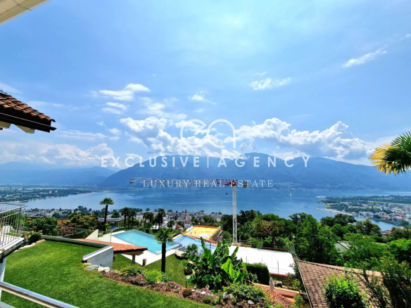 Panoramic view of a lake with mountains in the distance, with a landscaped garden and pool in the foreground on a sunny day