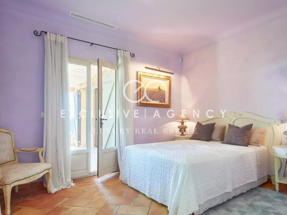 Pastel purple bedroom with white bedspread, cream headboard, and matching nightstands; glass doors open to a balcony with white curtains.