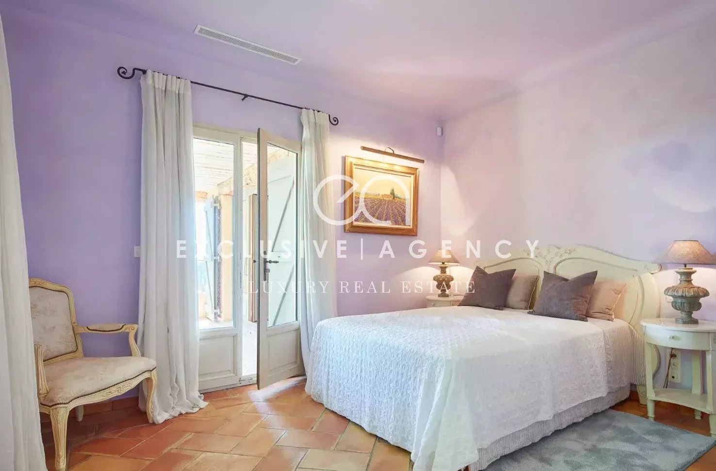 Pastel purple bedroom with white bedspread, cream headboard, and matching nightstands; glass doors open to a balcony with white curtains.