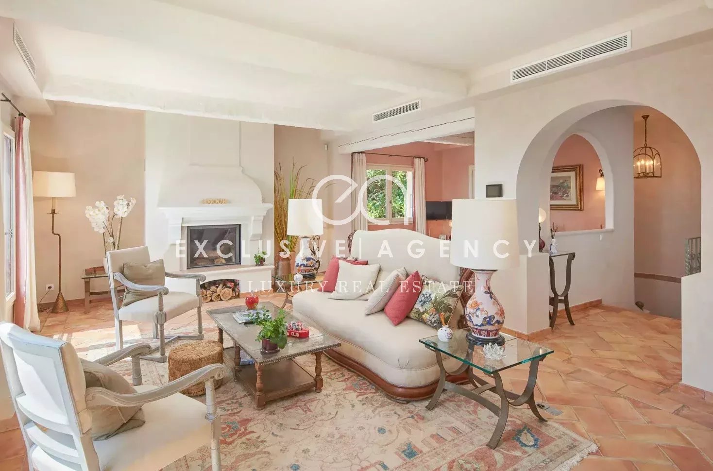 Luxurious living room with cream sofas and white armchairs around a wooden coffee table, facing a large white fireplace and terracotta tile floor.
