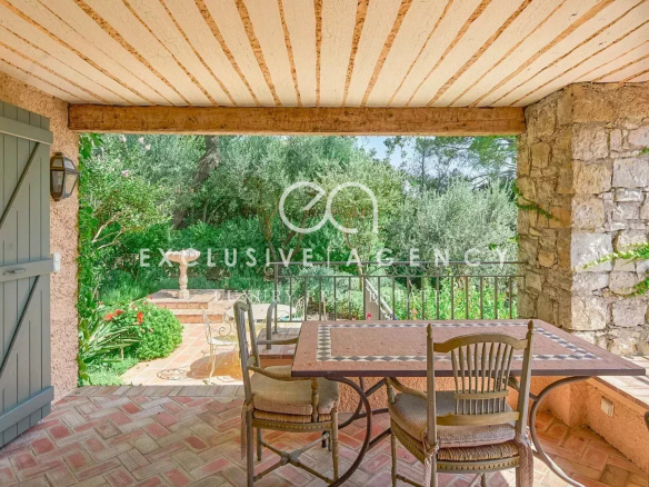 Covered patio with brick floor and wooden beams, patio table and chairs, open view to lush garden and stone wall terrace.