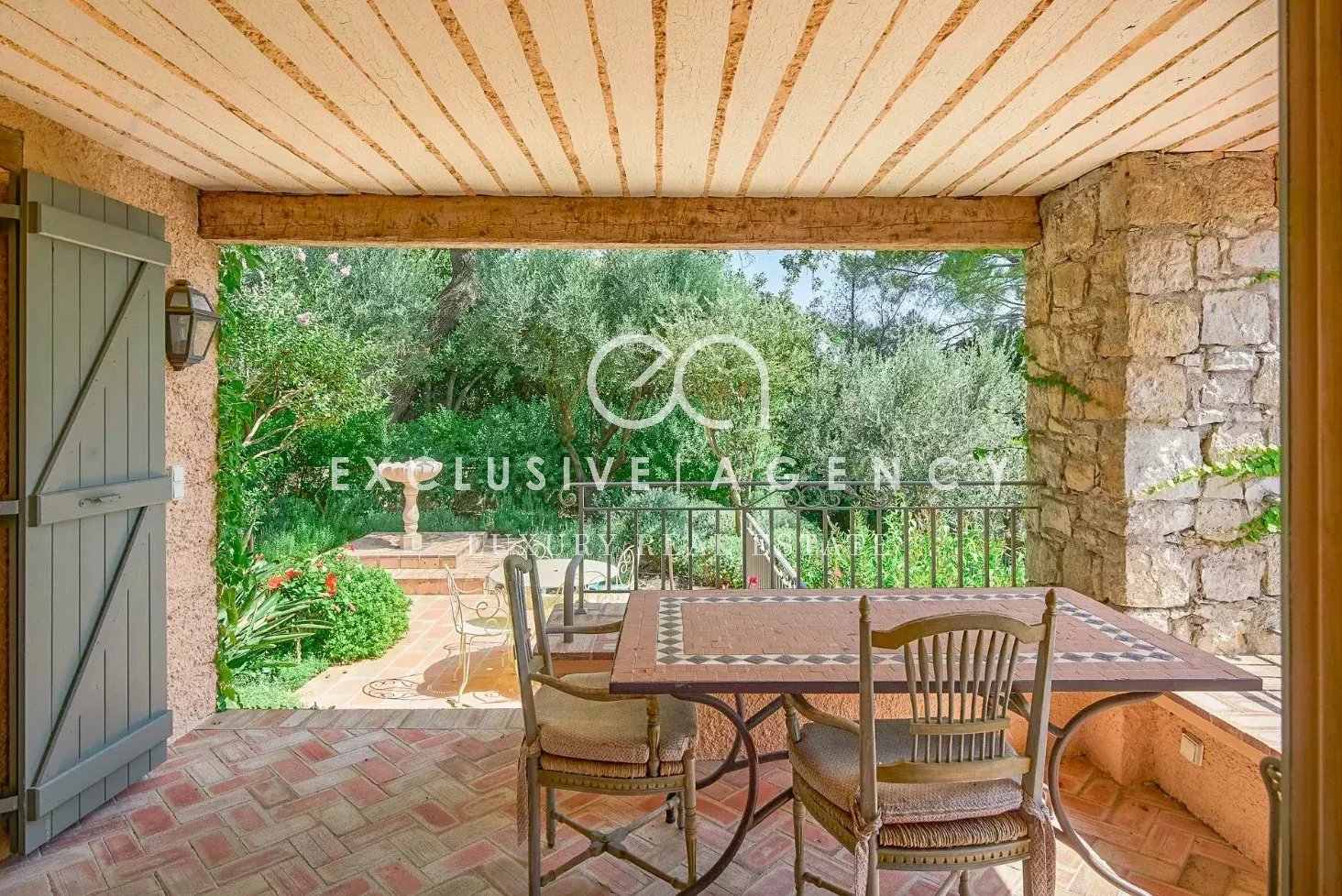 Covered patio with brick floor and wooden beams, patio table and chairs, open view to lush garden and stone wall terrace.