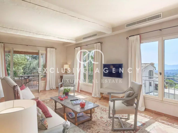 Bright living room with white furniture, cream curtains, and glass doors opening to a balcony with distant hills visible outside.