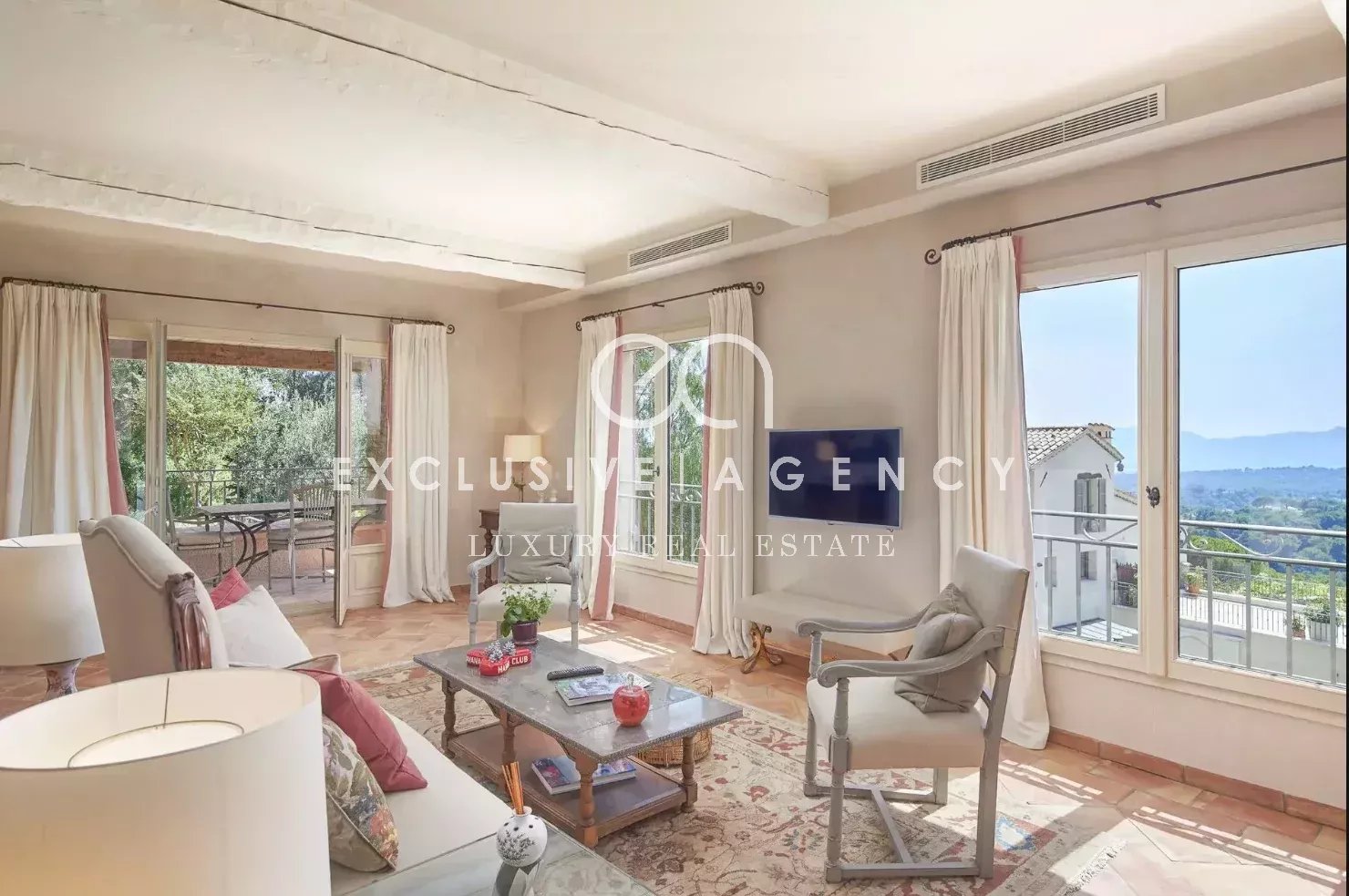 Bright living room with white furniture, cream curtains, and glass doors opening to a balcony with distant hills visible outside.