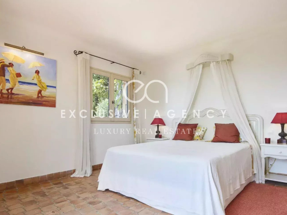 Bright white bedroom with a canopy bed, sheer curtains, and terracotta tiled floor. Two white nightstands with red lamps flank the bed. a beach painting hangs on the wall to the left.