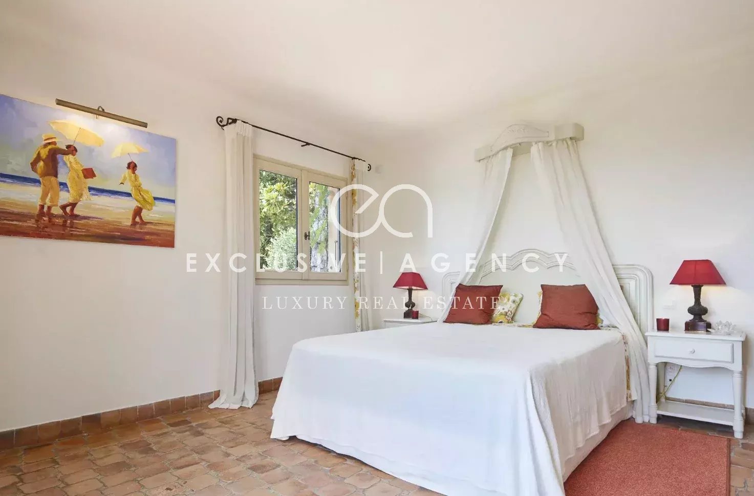 Bright white bedroom with a canopy bed, sheer curtains, and terracotta tiled floor. Two white nightstands with red lamps flank the bed. a beach painting hangs on the wall to the left.