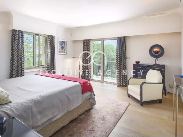 Luxurious bedroom with a king bed, gray bedding and red throw, facing large windows with patterned curtains and a balcony door.