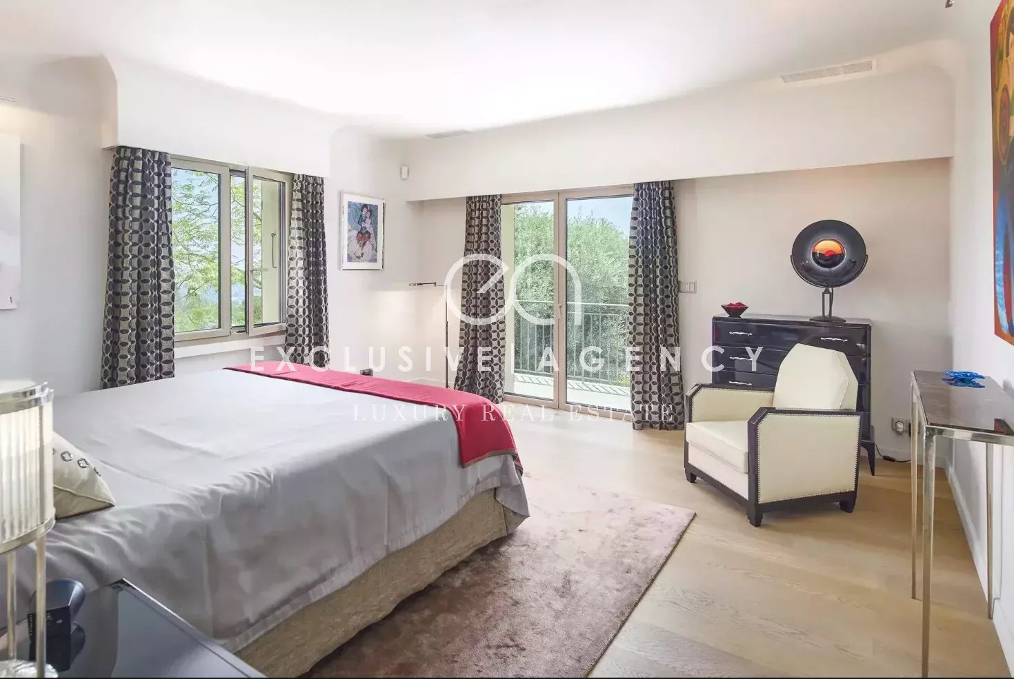Luxurious bedroom with a king bed, gray bedding and red throw, facing large windows with patterned curtains and a balcony door.
