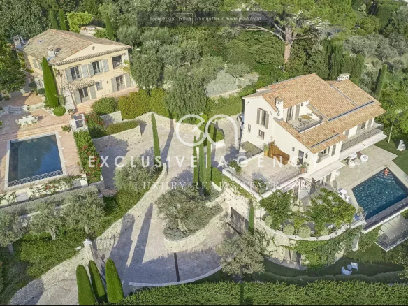Aerial view of a luxurious villa complex with multiple beige houses, terracotta roofs, manicured gardens, and a rectangular swimming pool surrounded by stone terraces.
