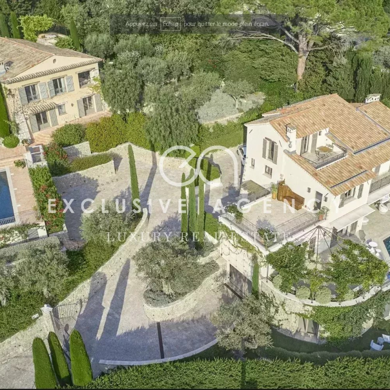 Aerial view of a luxurious villa complex with multiple beige houses, terracotta roofs, manicured gardens, and a rectangular swimming pool surrounded by stone terraces.