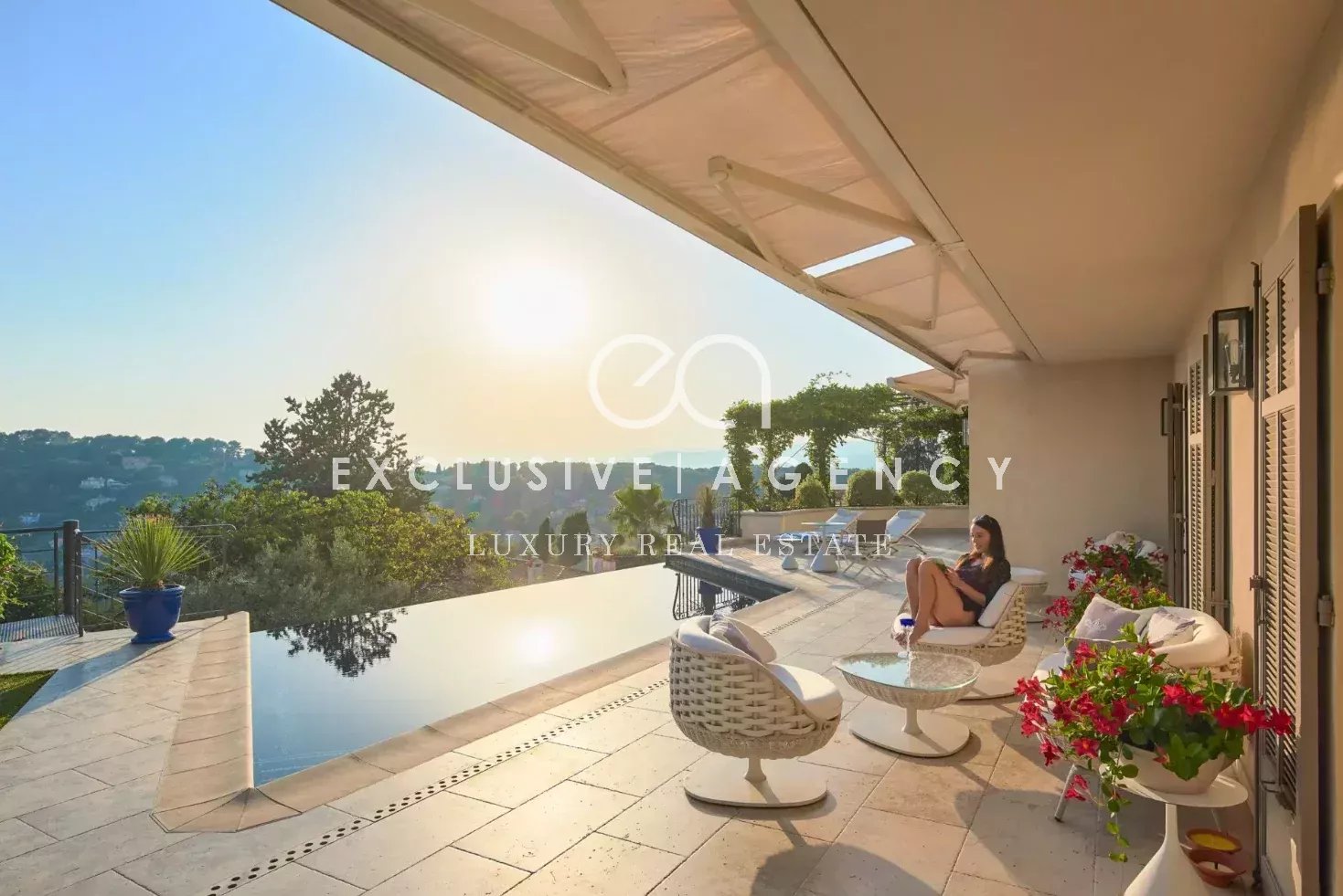 Luxurious outdoor terrace with an infinity pool, white wicker seating, and a woman relaxing in the sun overlooking hills at sunset.