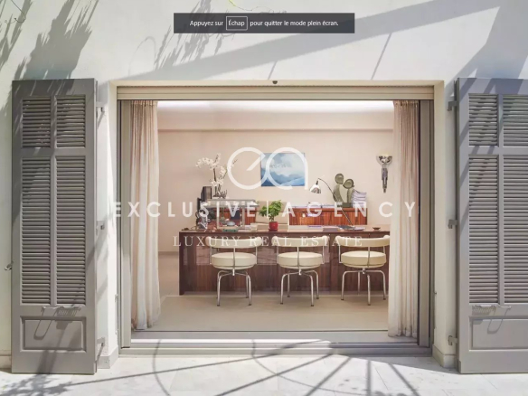 Open French doors with gray shutters reveal a luxurious dining room: long wooden table, cream chairs, plants, and soft curtains inside.