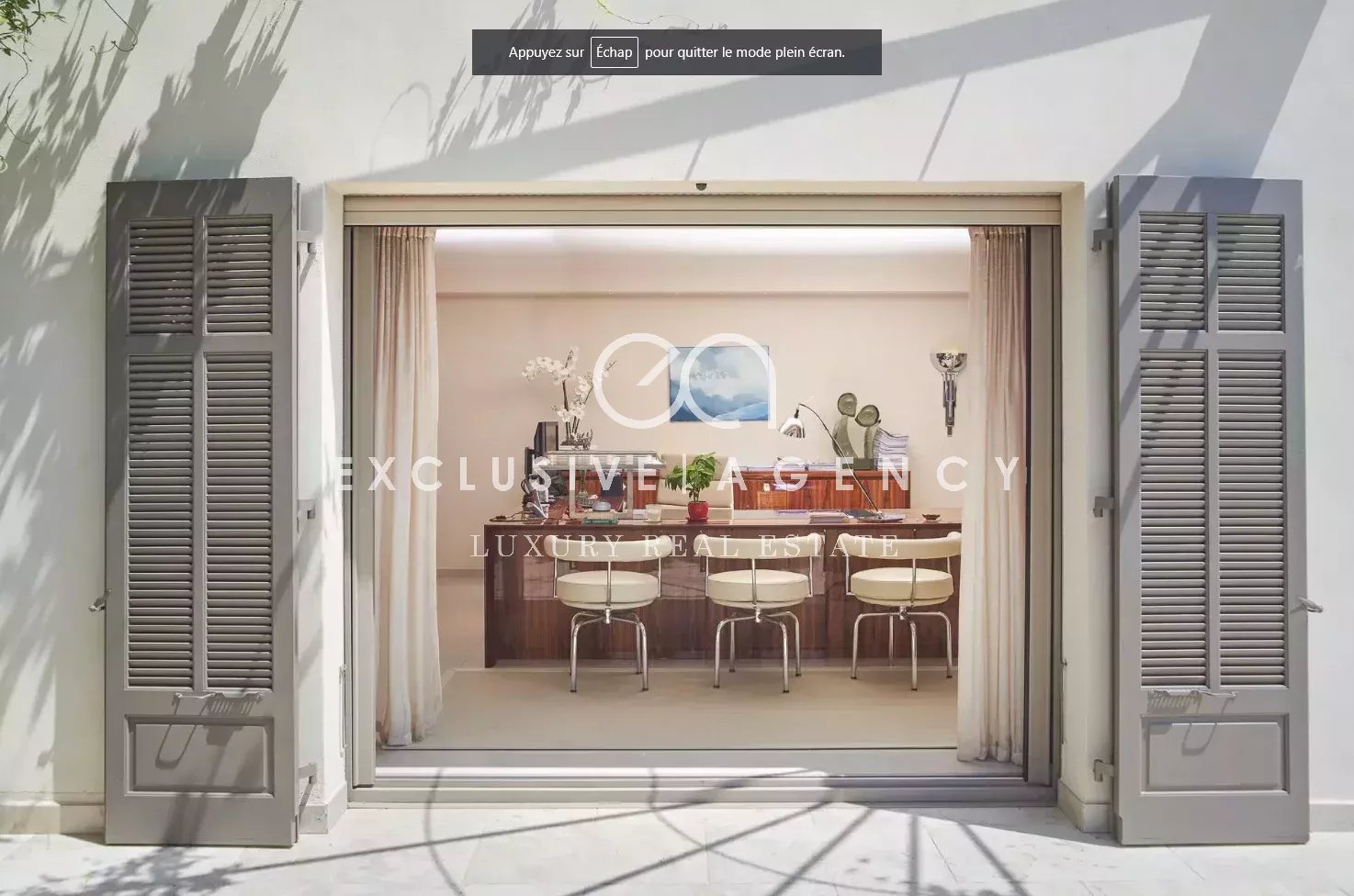 Open French doors with gray shutters reveal a luxurious dining room: long wooden table, cream chairs, plants, and soft curtains inside.