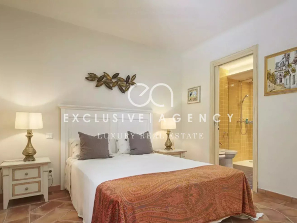 Bedroom with a white-paneled bed, two matching nightstands, warm lamps, and a red patterned throw at the foot of the bed; bathroom doorway nearby.