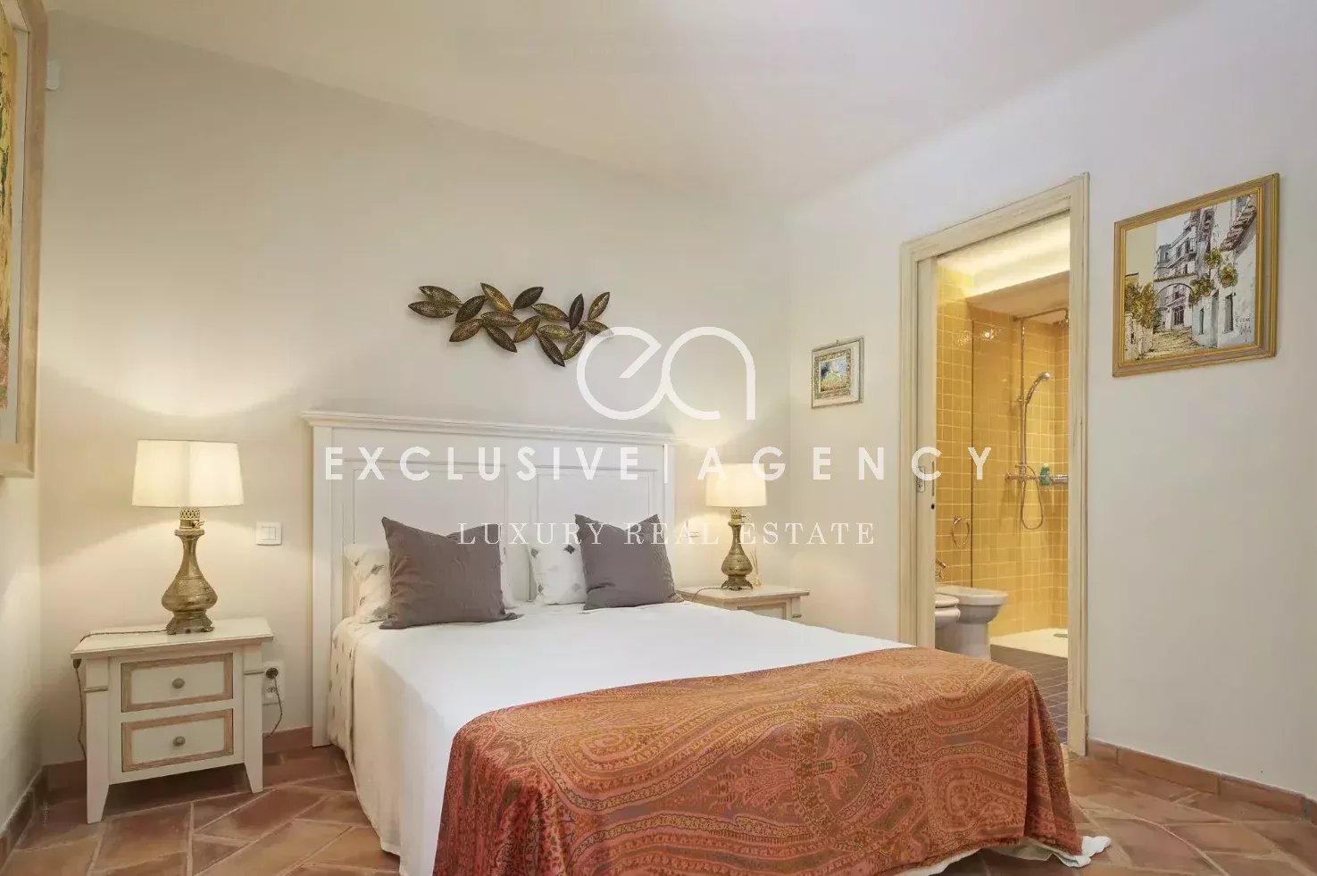 Bedroom with a white-paneled bed, two matching nightstands, warm lamps, and a red patterned throw at the foot of the bed; bathroom doorway nearby.