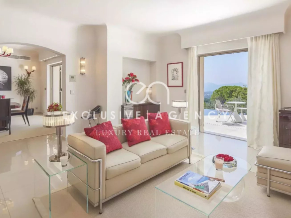Bright living room with white sofa and red cushions, glass coffee table, and a balcony with hillside view