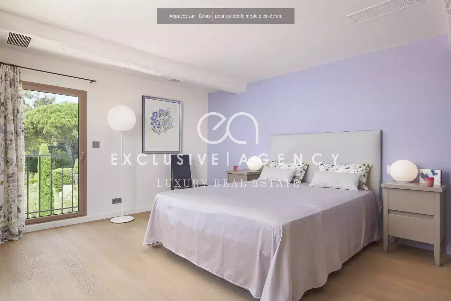 Bedroom with lavender accent wall and a large bed, two nightstands with globe lamps, and a balcony door opening to greenery.