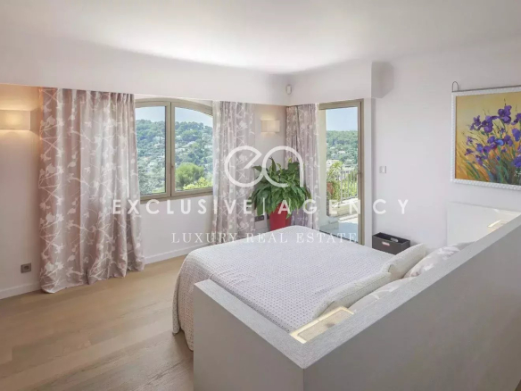 Bright bedroom with a low white platform bed, gray bedding, and large windows overlooking a hillside, framed by pink floral curtains.