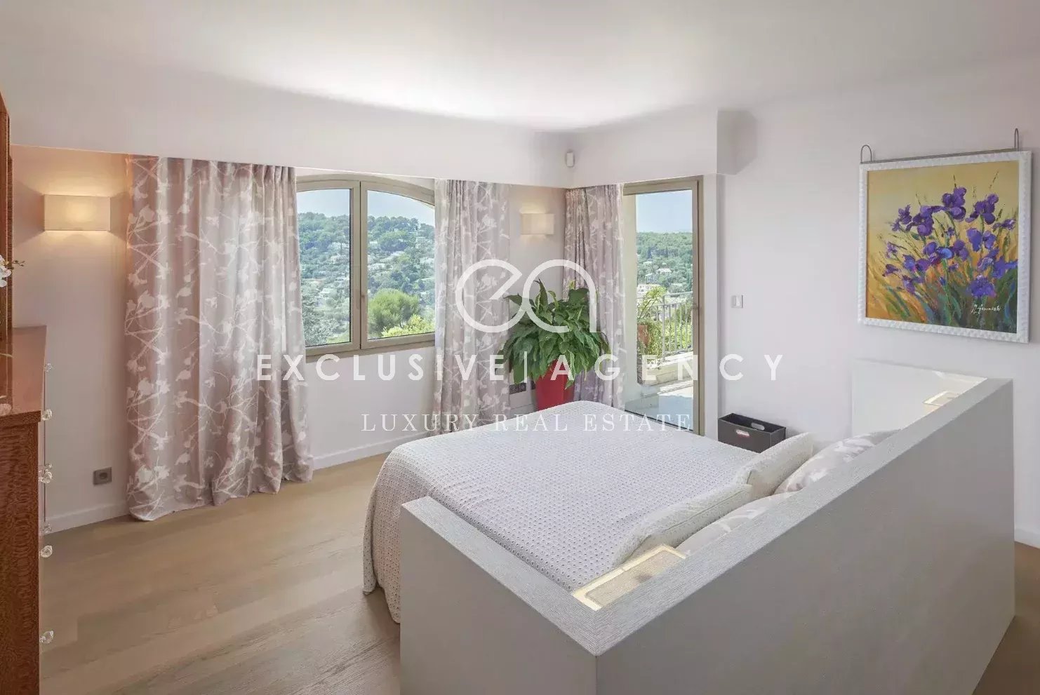 Bright bedroom with a low white platform bed, gray bedding, and large windows overlooking a hillside, framed by pink floral curtains.