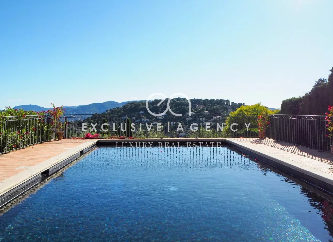Rooftop swimming pool with blue water, metal railing, and potted plants, overlooking green hills under a clear sky.