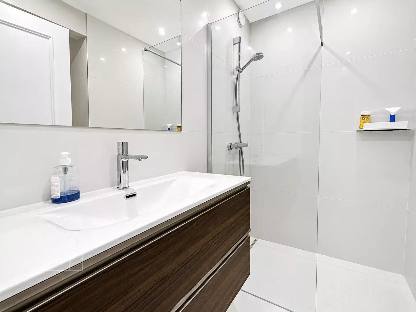 Salle de bain moderne avec douche, lavabo et miroir, murs et sol en carrelage clair.