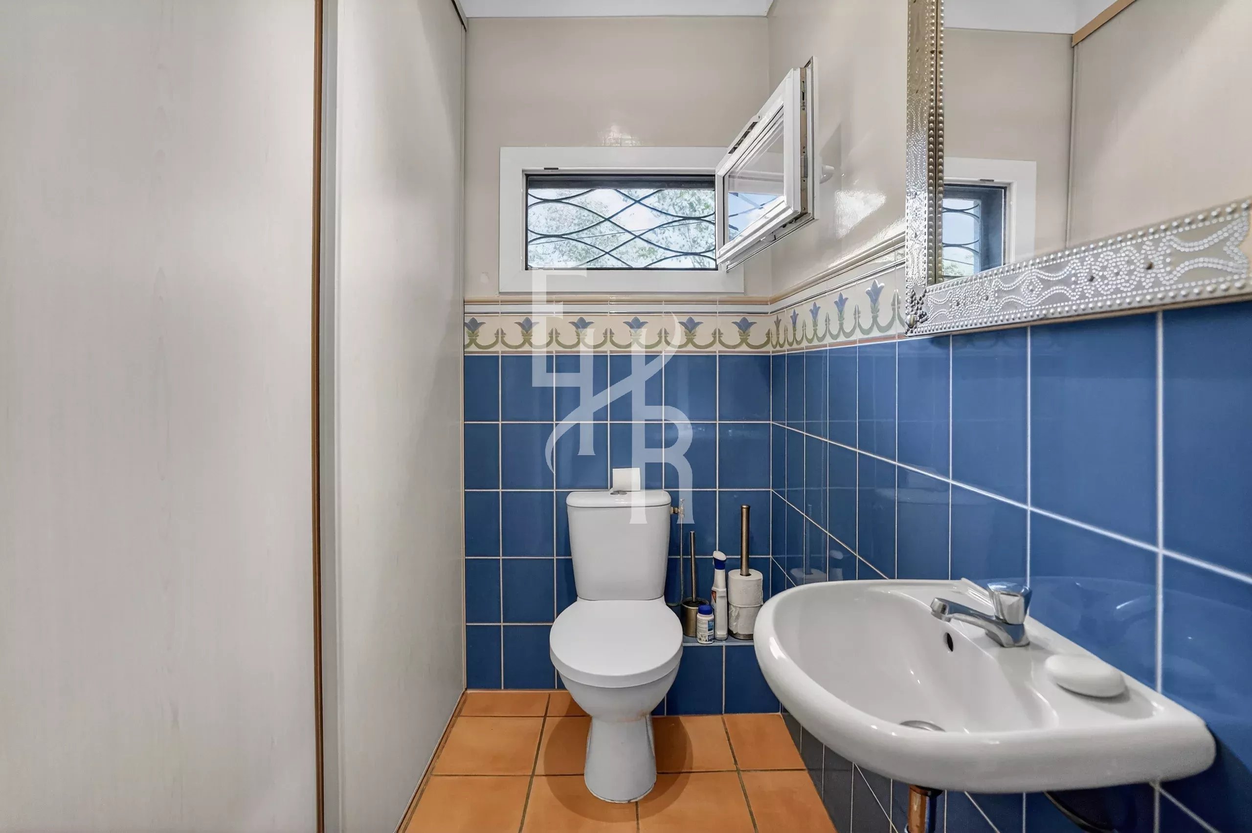 Compact bathroom with blue tile walls, orange floor, and white toilet and sink; small window above the toilet is open.
