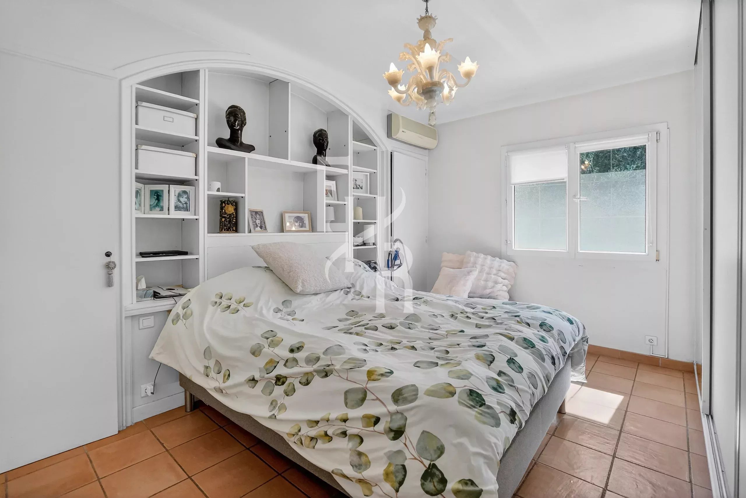 White bedroom with built-in shelving behind the bed, bust sculptures, framed photos, and a botanical duvet on a low bed.