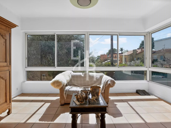 Bright sunlit sunroom with a wicker loveseat covered in a beige throw, a wooden coffee table with teapots, and a tall wooden cabinet on the left; large windows overlook houses outside.