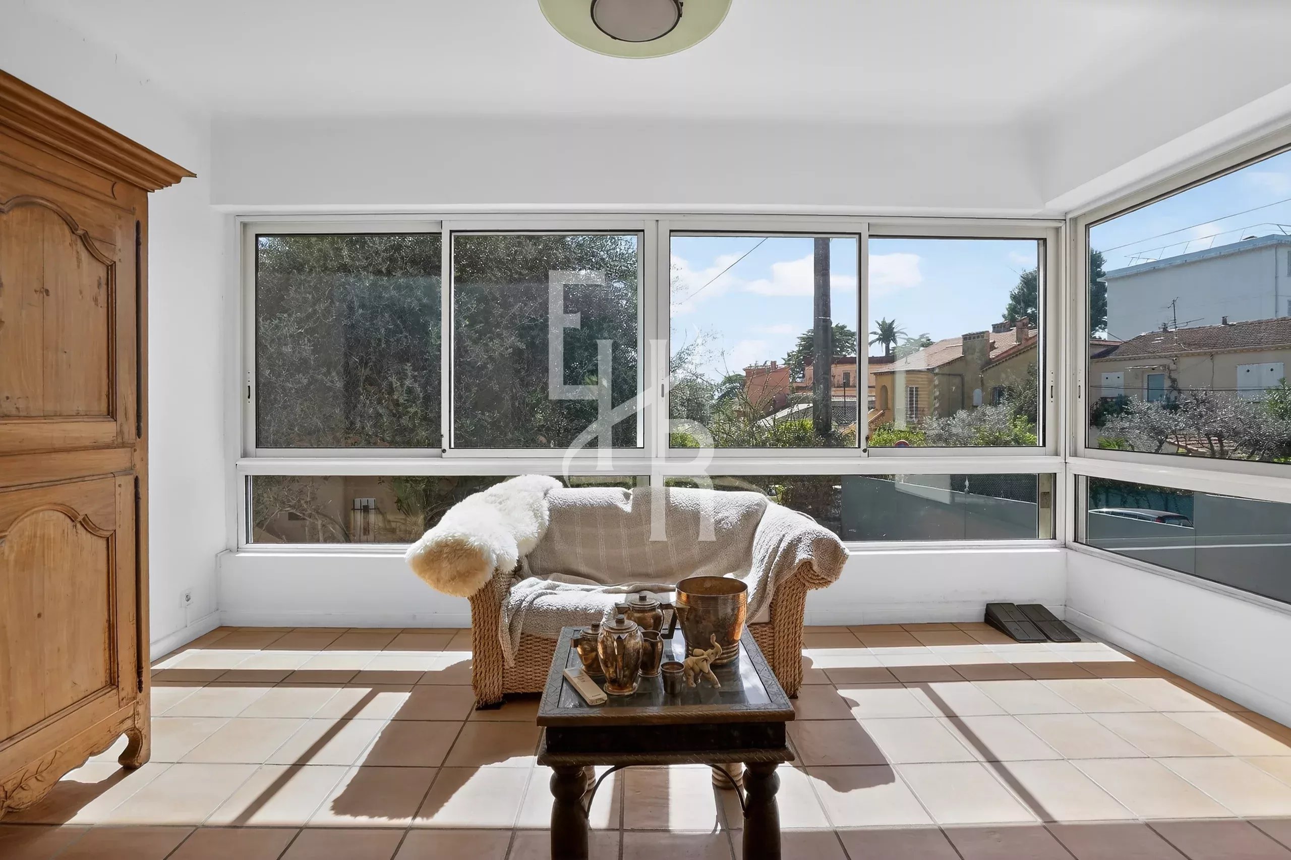 Bright sunlit sunroom with a wicker loveseat covered in a beige throw, a wooden coffee table with teapots, and a tall wooden cabinet on the left; large windows overlook houses outside.