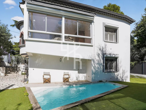 White two-story house with a large glass-enclosed room on the upper level, a rectangular pool in the yard, and two wooden deck chairs on the patio.