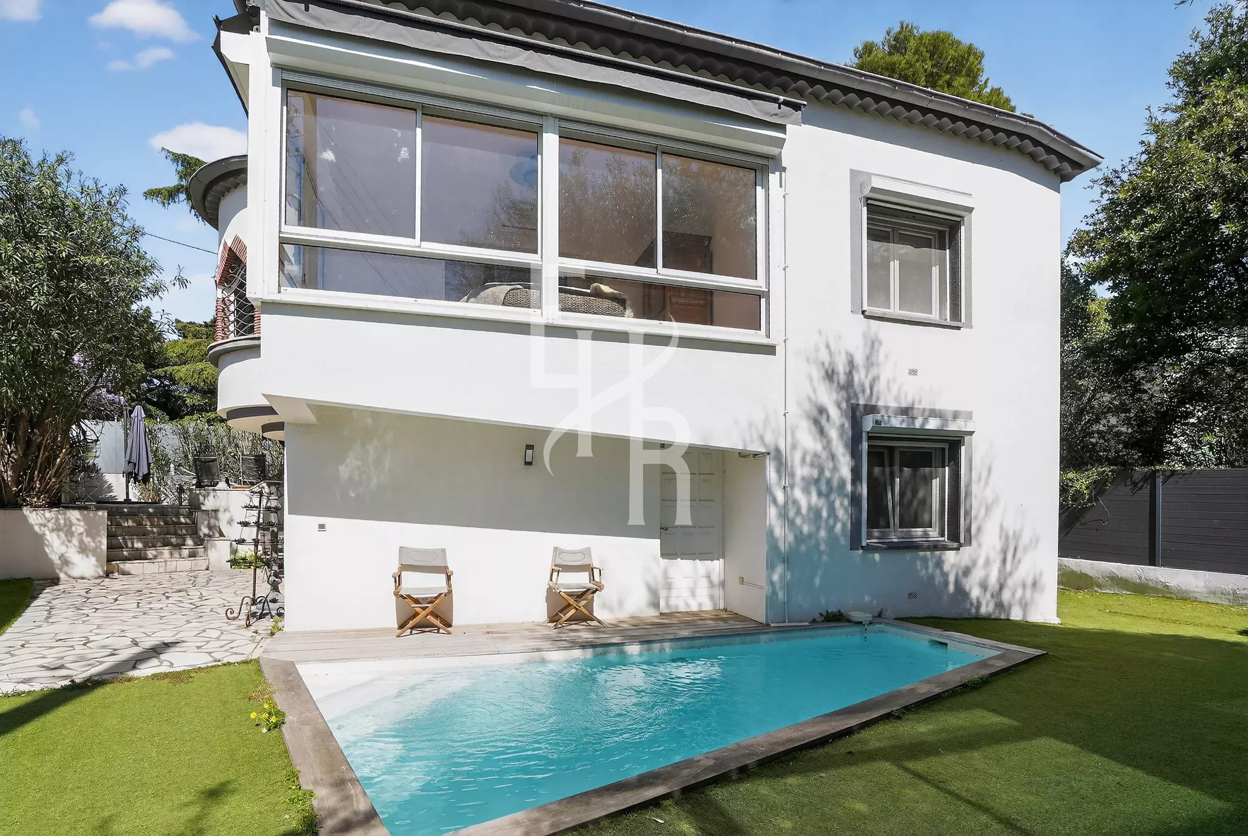 White two-story house with a large glass-enclosed room on the upper level, a rectangular pool in the yard, and two wooden deck chairs on the patio.