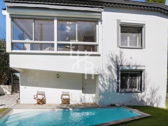 White two-story house with large glass windows, a rectangular pool in the yard, and two wooden deck chairs on a wooden deck by the pool.