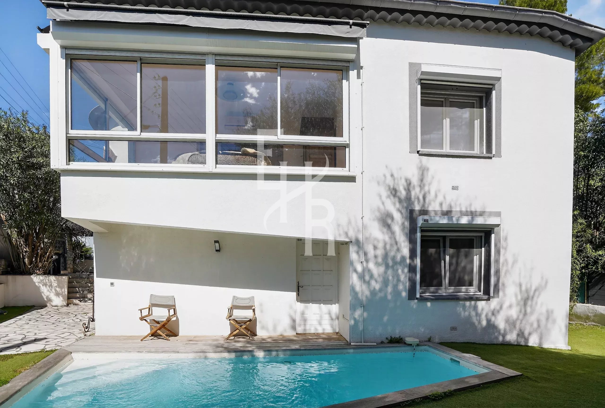 White two-story house with large glass windows, a rectangular pool in the yard, and two wooden deck chairs on a wooden deck by the pool.