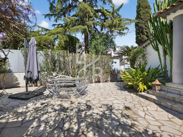 Stone-paved outdoor patio with a closed umbrella, metal table and chairs, and surrounding greenery under a blue sky.