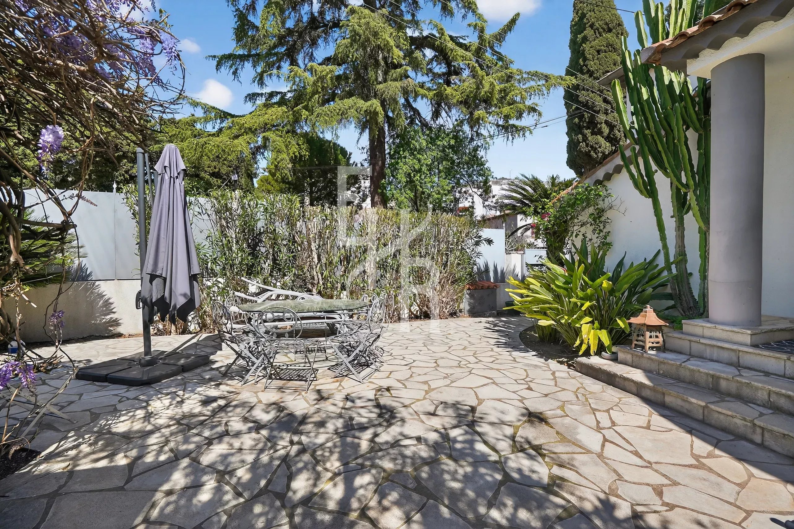 Stone-paved outdoor patio with a closed umbrella, metal table and chairs, and surrounding greenery under a blue sky.
