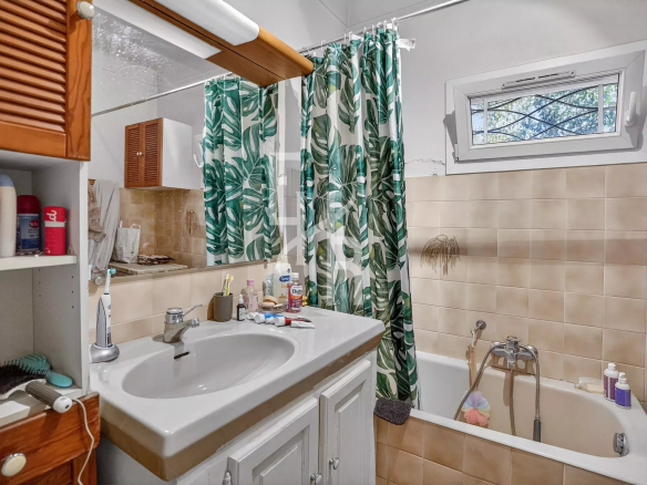 Bathroom with beige-tiled walls, white sink and cabinet, and a green tropical leaf shower curtain beside a bathtub under a small window.