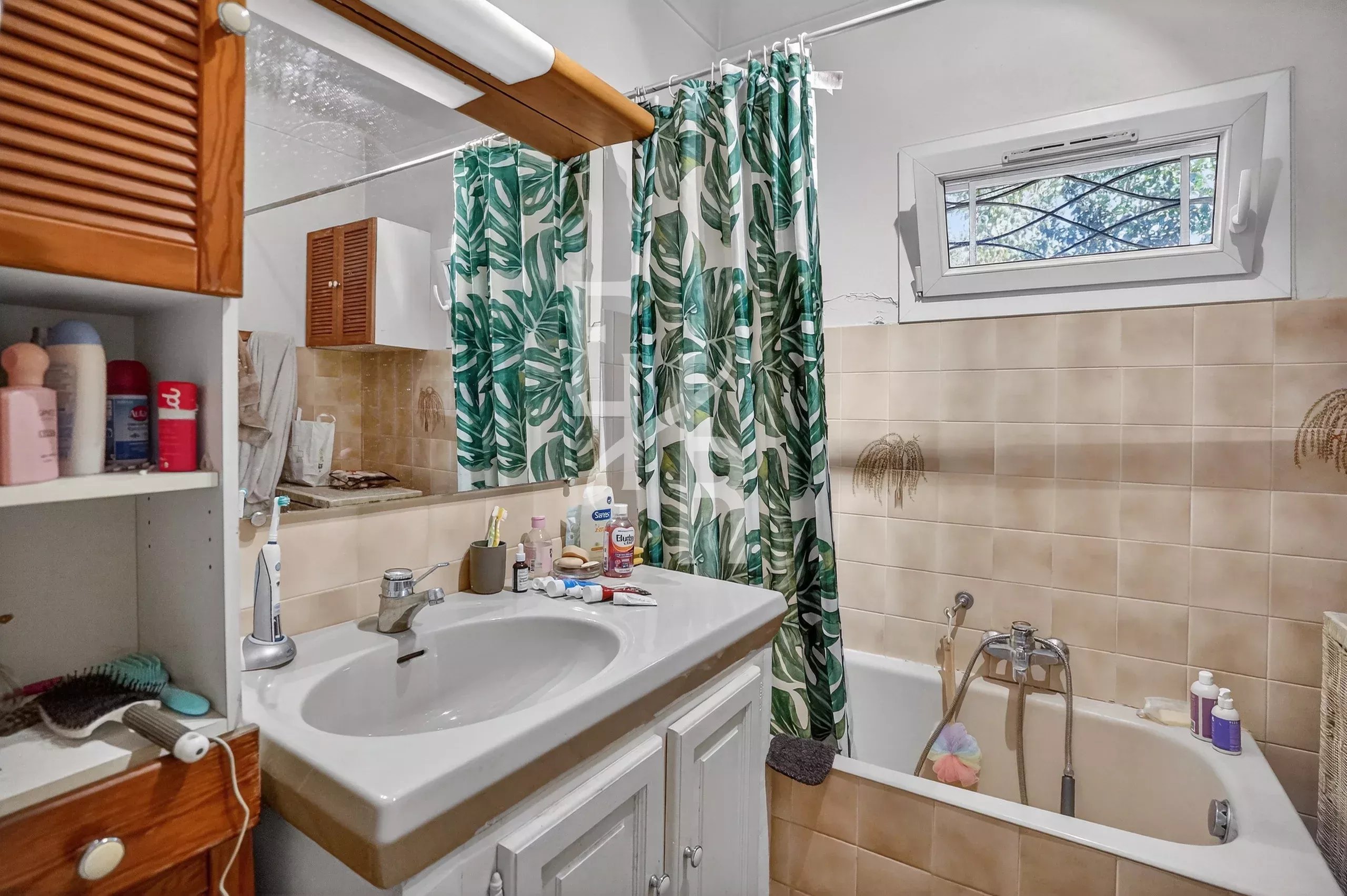 Bathroom with beige-tiled walls, white sink and cabinet, and a green tropical leaf shower curtain beside a bathtub under a small window.