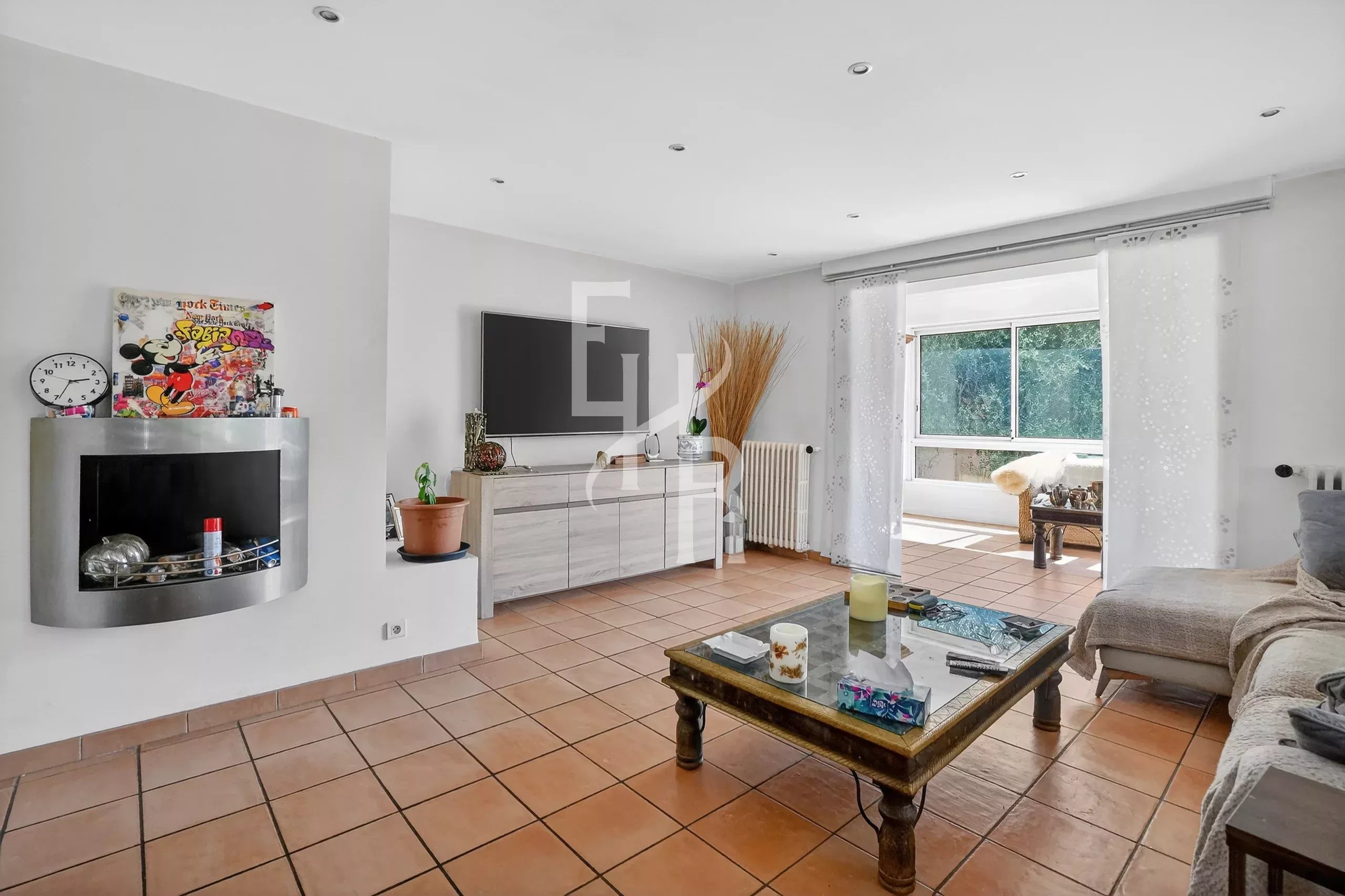 Bright living room with a wall-mounted flat-screen TV over a white cabinet, a fireplace to the left, and terracotta tile flooring under a glass coffee table and sofa setup.