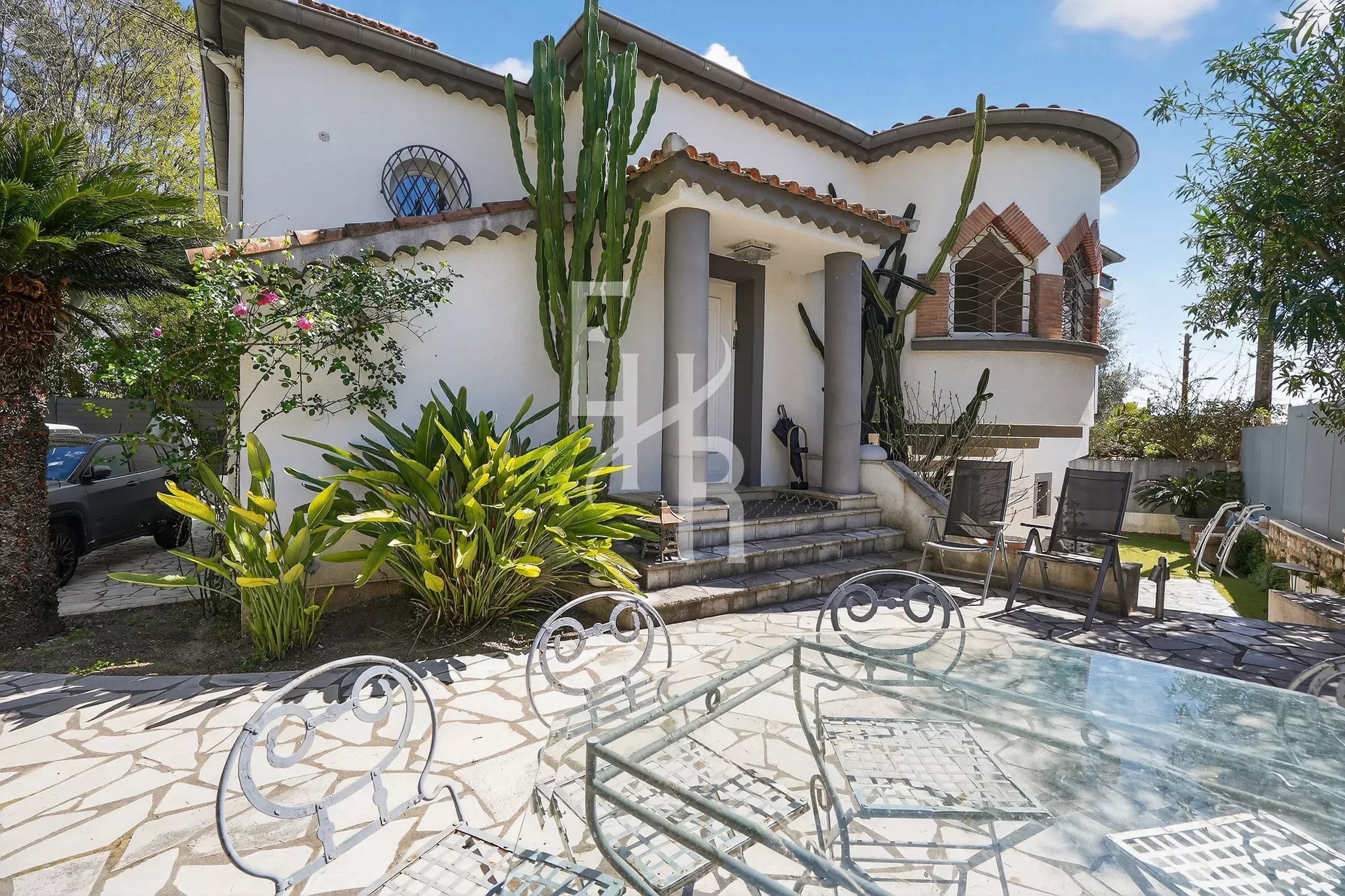 White house with rounded tower and tall cacti at the entrance, stone steps, and a glass-top patio table with iron chairs in a sunny backyard.