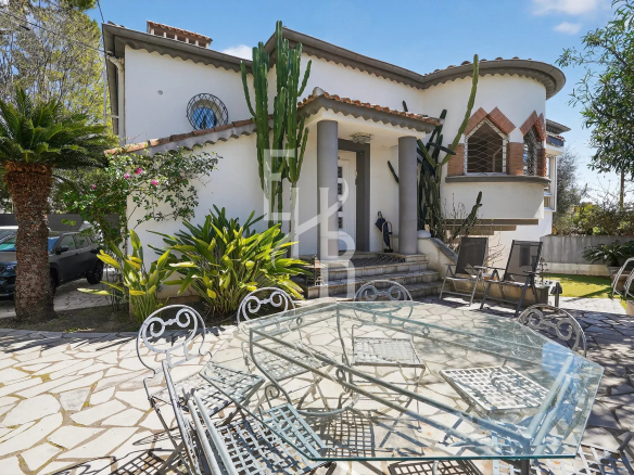 White stucco house with curved rooflines, tall cacti by the entrance, and a glass patio table with metal chairs on a stone courtyard.