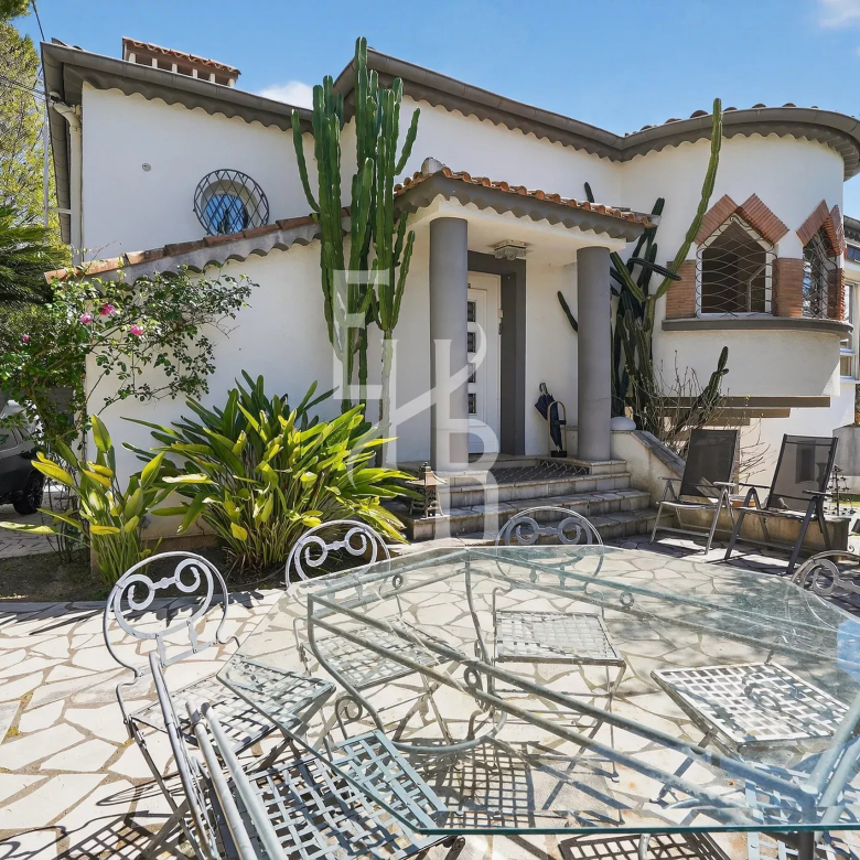 White stucco house with curved rooflines, tall cacti by the entrance, and a glass patio table with metal chairs on a stone courtyard.