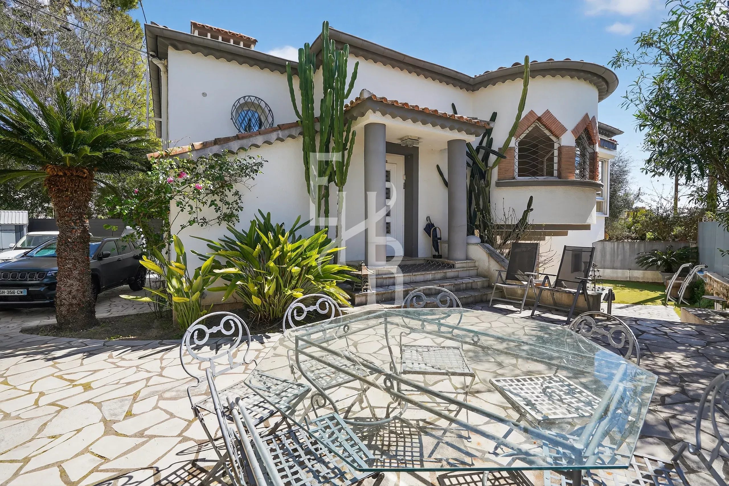 White stucco house with curved rooflines, tall cacti by the entrance, and a glass patio table with metal chairs on a stone courtyard.