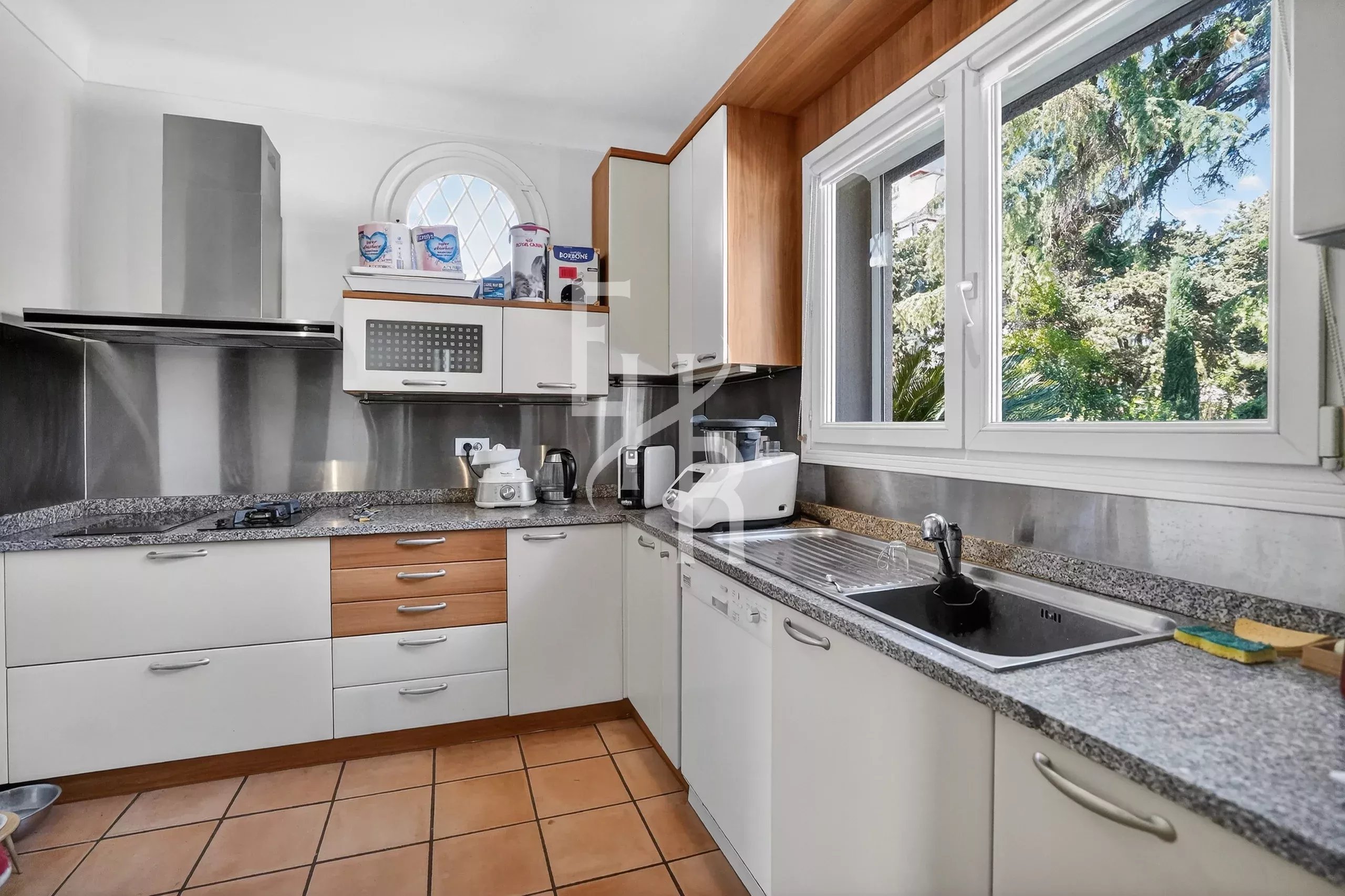 Bright L-shaped kitchen with white cabinets, granite countertops, and a large window overlooking greenery; sink and appliances along the counter.