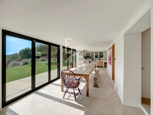 Interior of a modern country house with a large wooden table and bay windows overlooking a garden.
